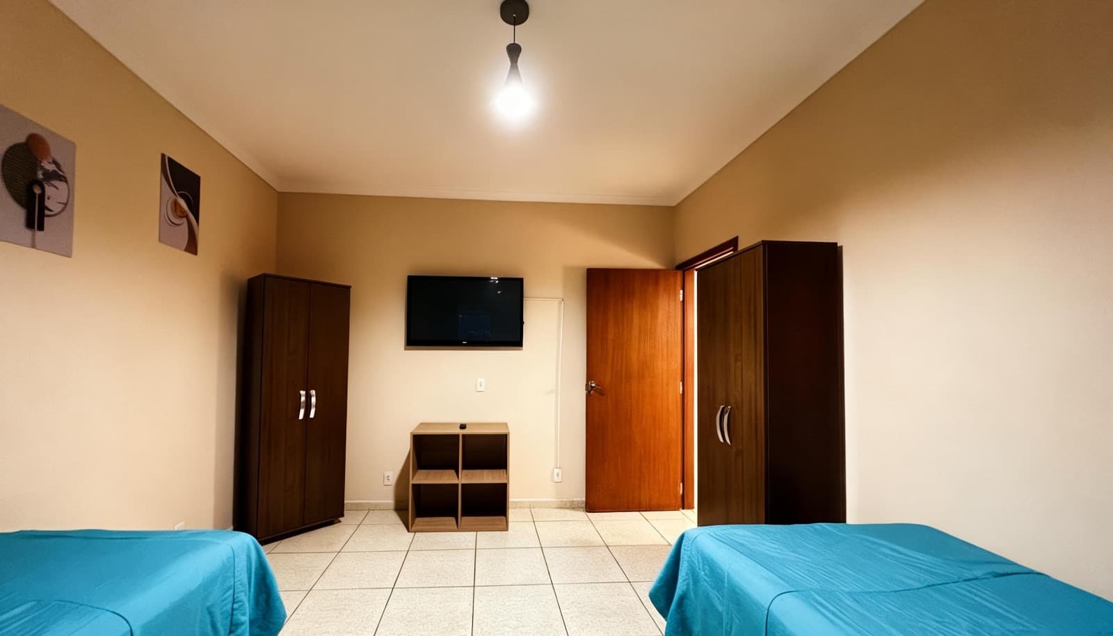 Quarto com duas camas, armários, televisão e área central de circulação.