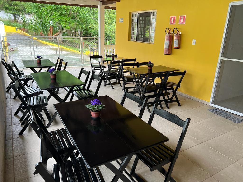 Refeitório com mesas e cadeiras sob varanda de parede amarela.