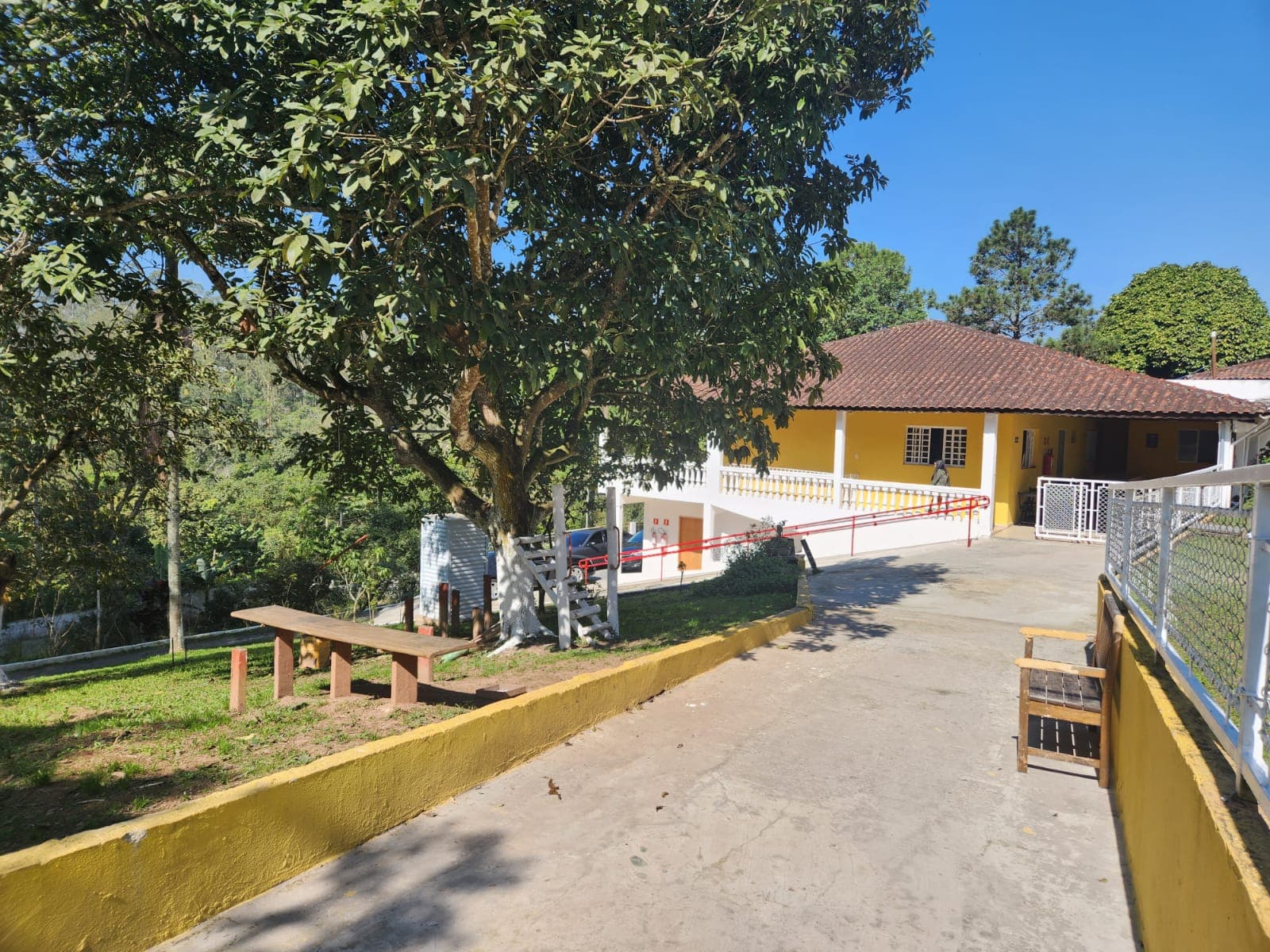Área externa da Arhat com árvores, banco, rampa e fachada amarela.