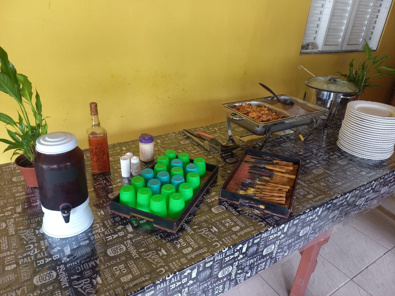 Mesa de refeição com bebidas, recipientes térmicos, pratos e utensílios.
